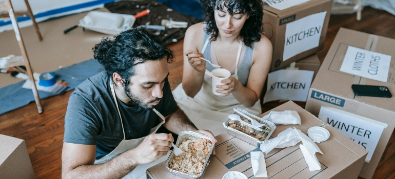 Couple sitting on the floor eating takeout food during a home move with labeled cardboard boxes around them.