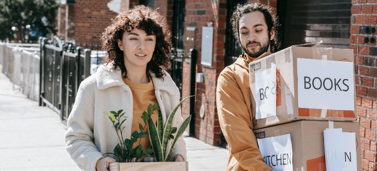 Man and woman walking outside with labeled moving boxes and a houseplant during their relocation.